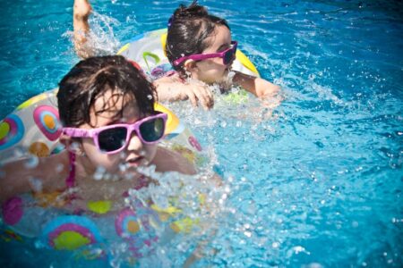 Two young girls in sunglasses and swim rings splash in a blue swimming pool on a sunny day.