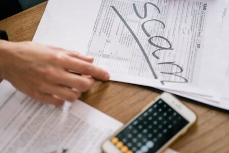 Hands hold a document marked 'SCAM' next to a smartphone with a calculator, symbolizing fraud prevention.