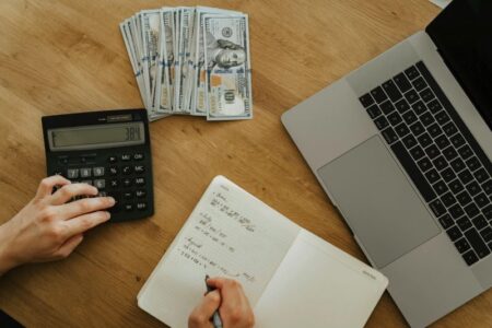 Hands using a calculator and writing in a notebook, with stacks of money and a laptop on a wooden desk.