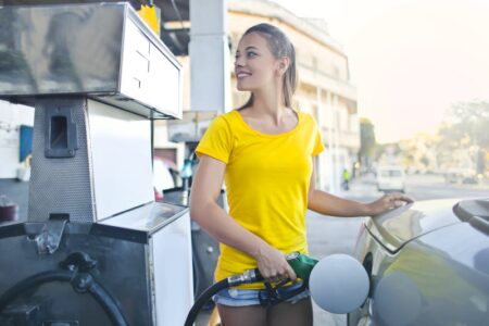 A smiling woman in a yellow shirt pumps gas into her car at a gas station.
