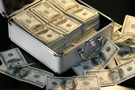 Briefcase overflowing with stacks of US $100 bills, with more bills scattered around on a dark surface.