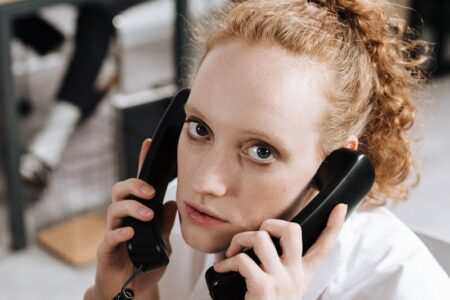 A person with red hair holds two black phone receivers to their ears, looking intently at the camera.
