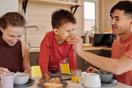 A father feeds his smiling young child a piece of fruit at a kitchen table, with the mother smiling nearby.