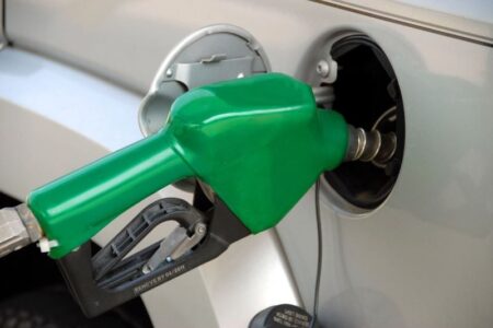 A green gas pump nozzle is inserted into the fuel tank of a silver car, illustrating fuel prices.