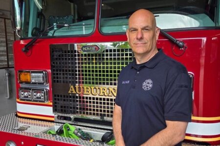 A bald man in a dark polo shirt stands in front of an Auburn Fire Department truck.
